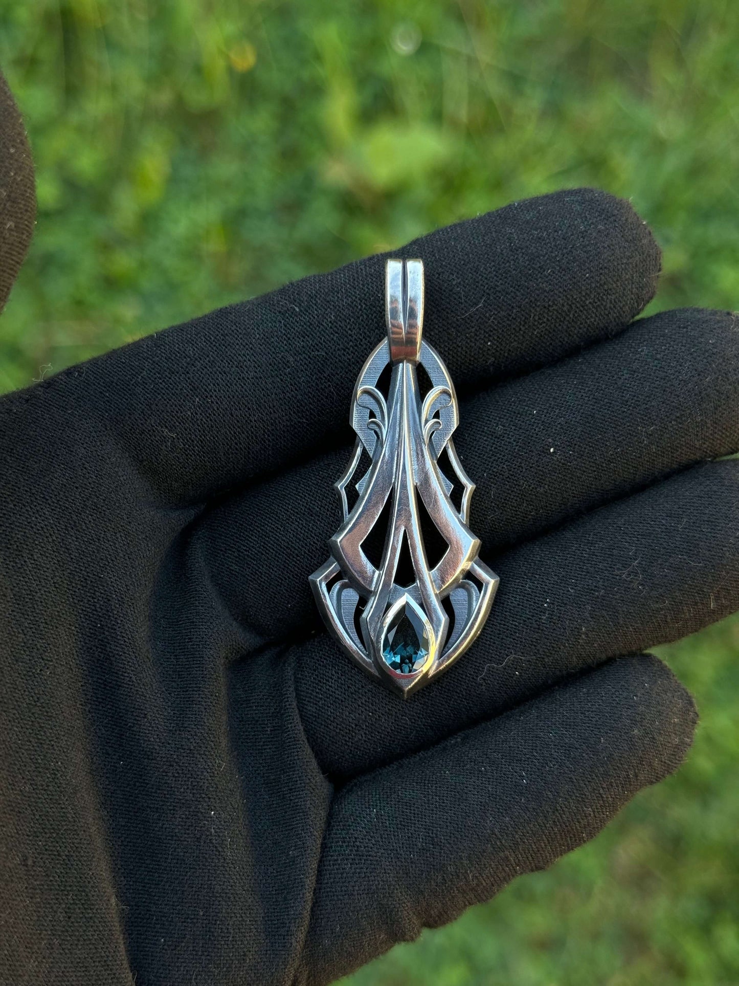 Silver pendant with an intricate design and a pear-shaped blue gemstone, held by a gloved hand against a grassy background