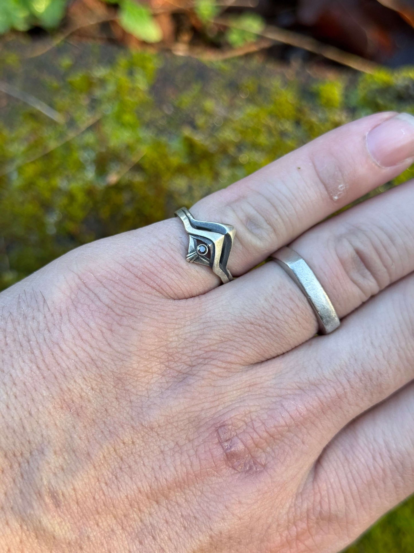 Hand wearing a Silver ring with a sleek, modern design and a small black gemstone, displayed on vibrant green moss
