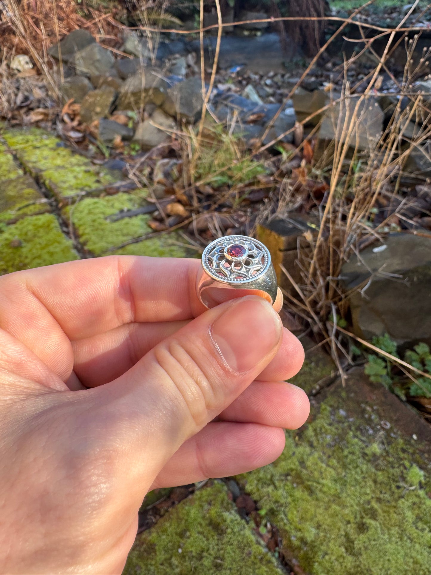 Hand holding a silver ring with a floral design and a red gemstone, set against a mossy outdoor scene with rocks and plants