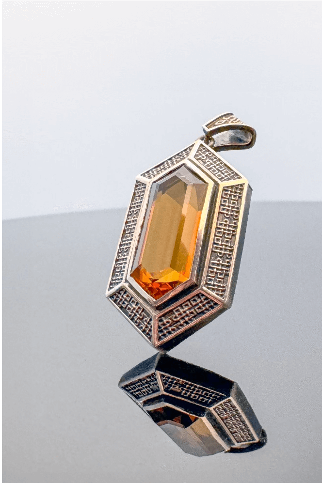 Hexagonal silver pendant with intricate patterns and a large orange gemstone, reflecting on a smooth surface
