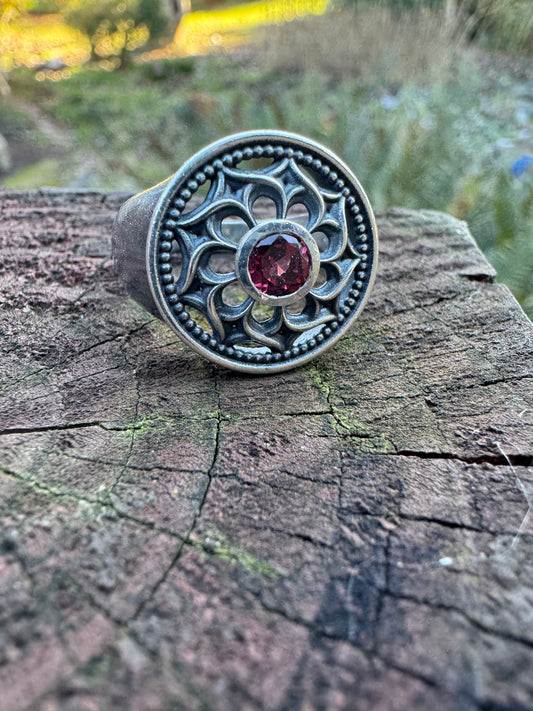 Silver ring with a floral design and a red gemstone displayed on a wood surface, set against a outdoor scene 