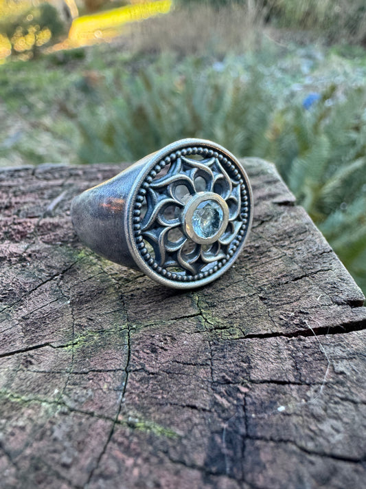 Silver ring with a floral design and a blue gemstone displayed on a wood surface, set against a outdoor scene