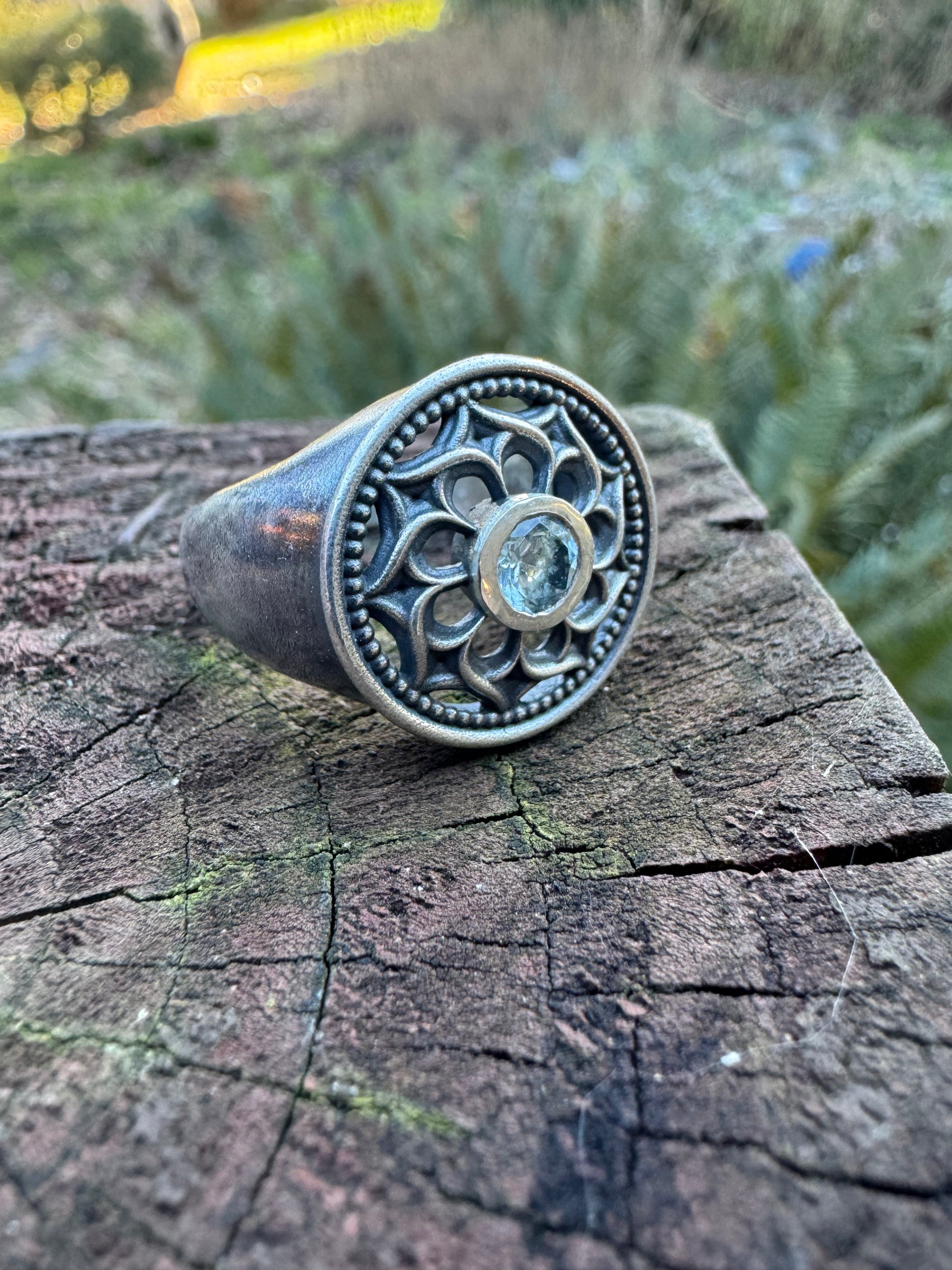 Silver ring with a floral design and a blue gemstone displayed on a wood surface, set against a outdoor scene