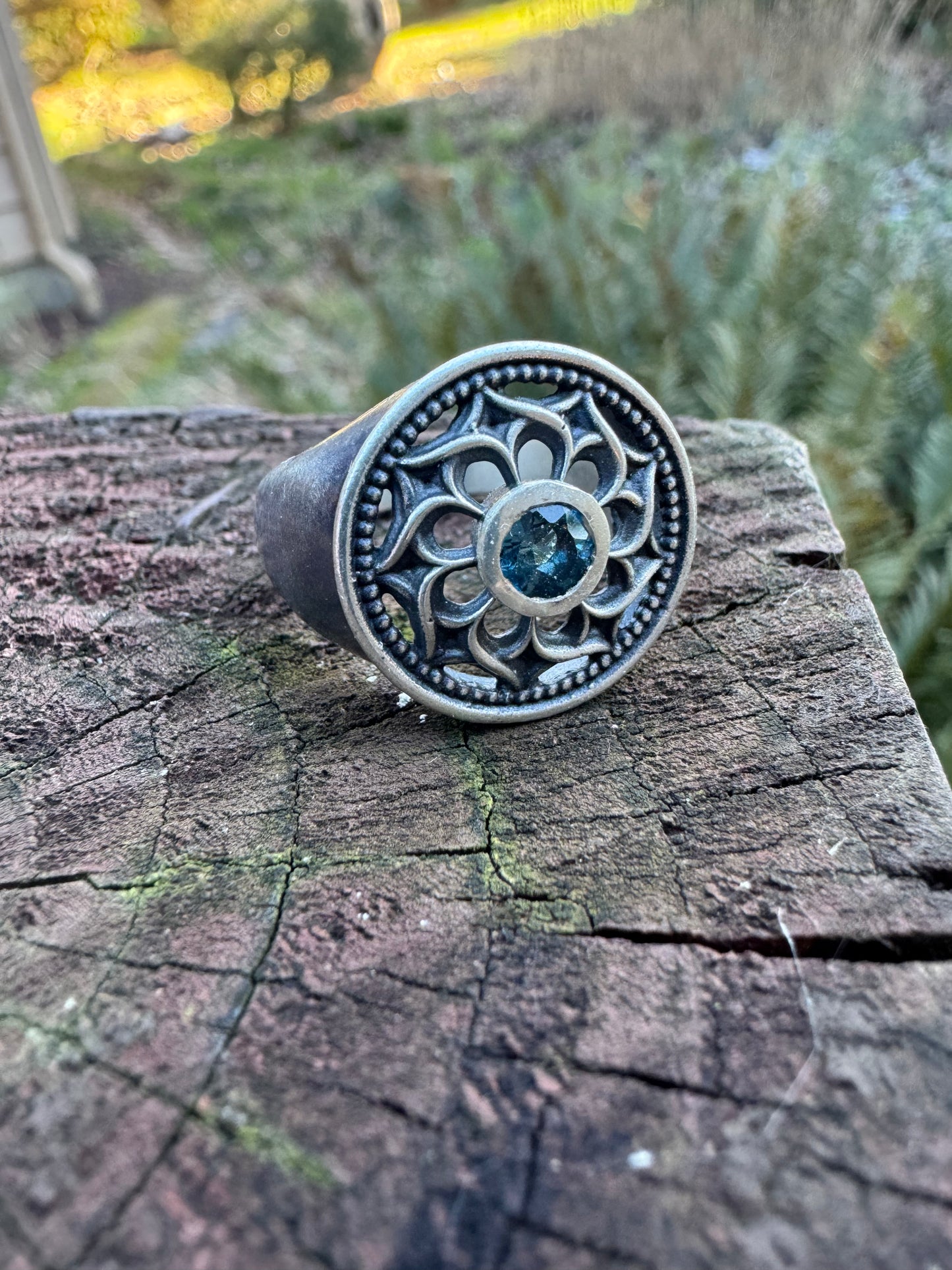 Silver ring with a floral design and a blue gemstone displayed on a wood surface, set against a outdoor scene