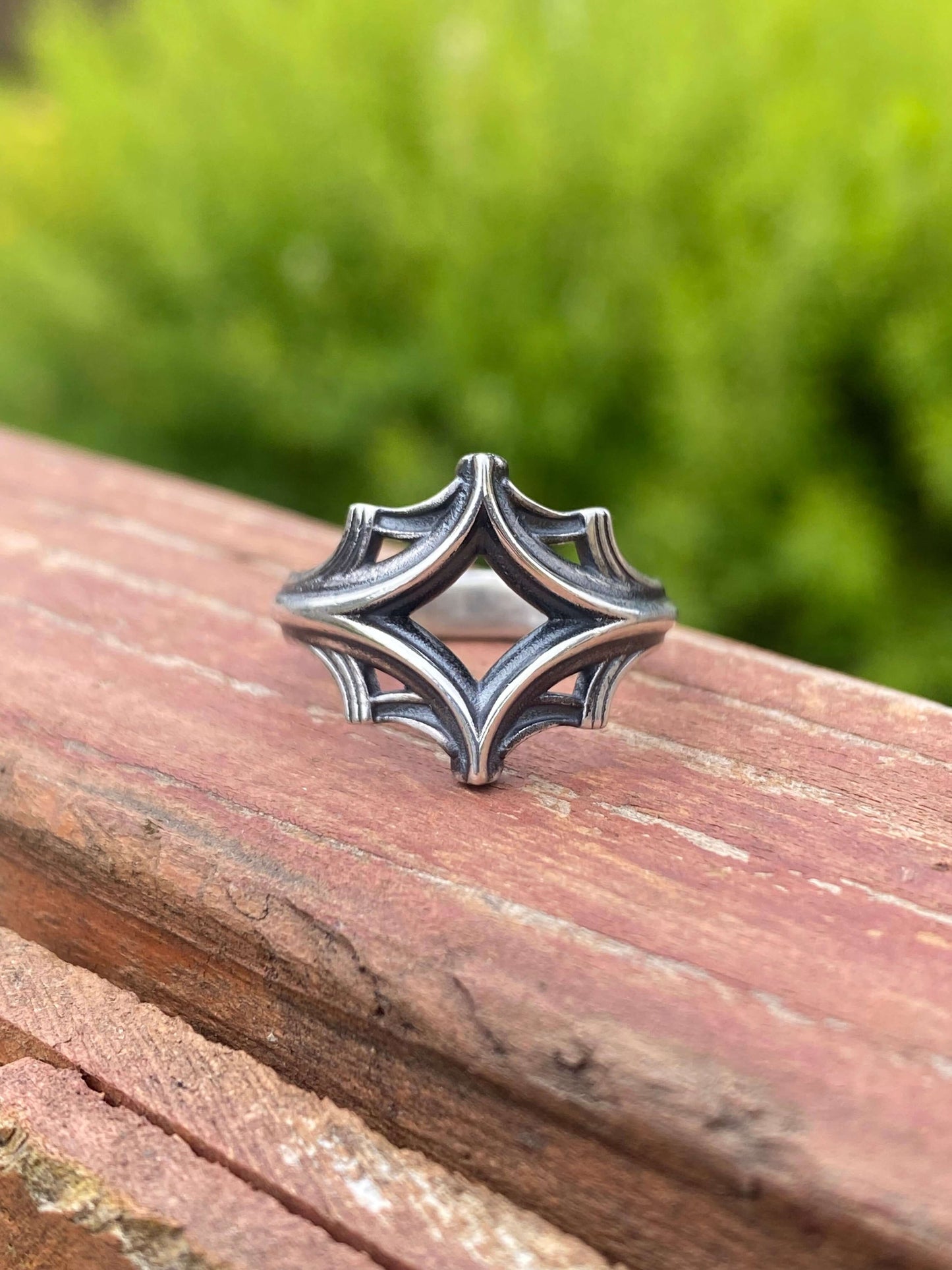 Silver ring with an intricate geometric design, displayed on a wooden surface with a blurred green background