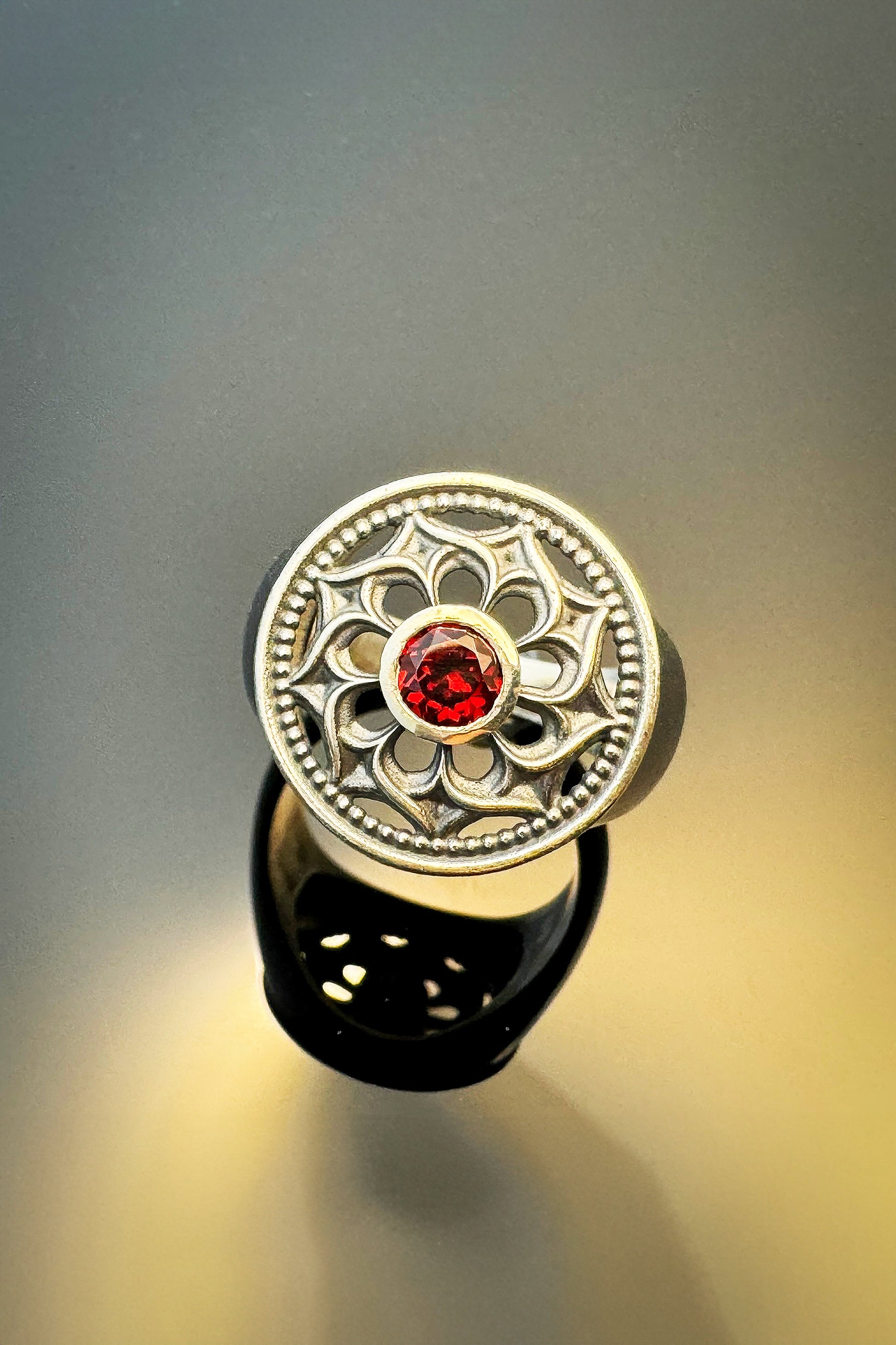 Silver ring featuring an ornate floral design with a central red gemstone, displayed against a soft gradient background