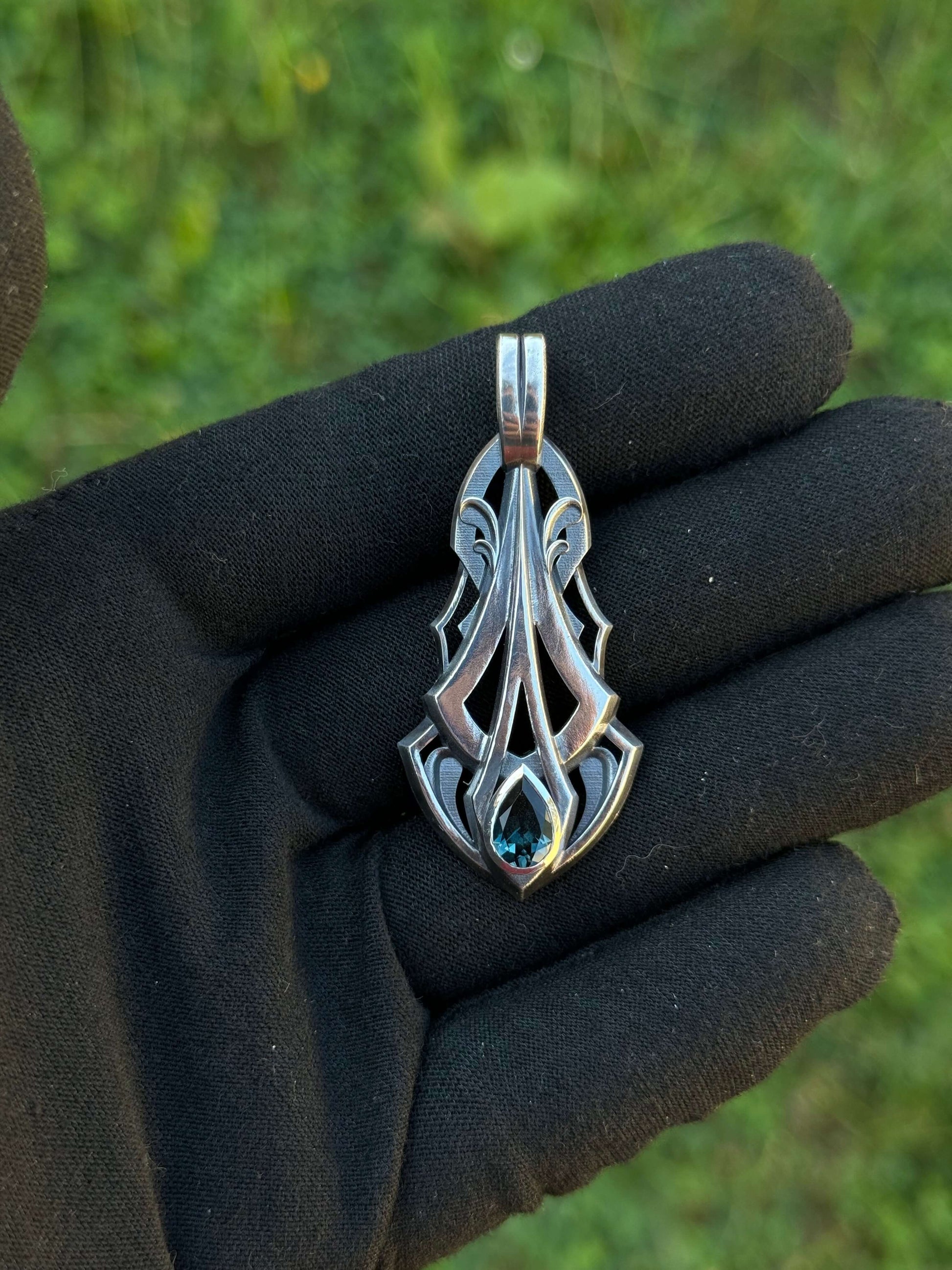 Silver pendant with an intricate design and a pear-shaped blue gemstone, held by a gloved hand against a grassy background