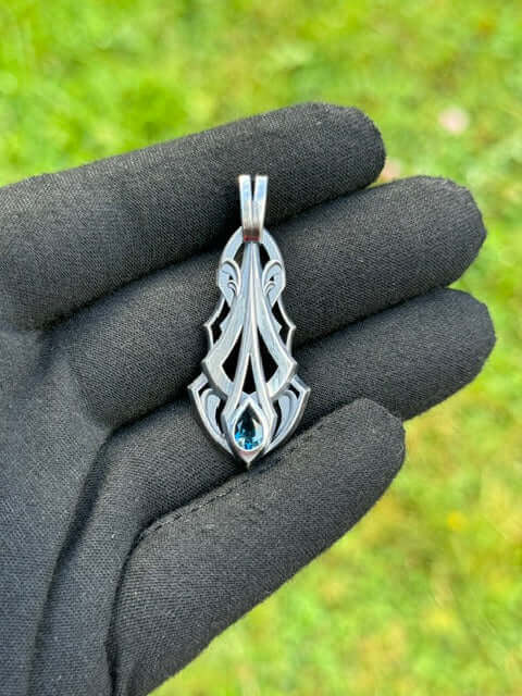 Silver pendant with an intricate design and a pear-shaped blue gemstone, held by a gloved hand against a grassy background