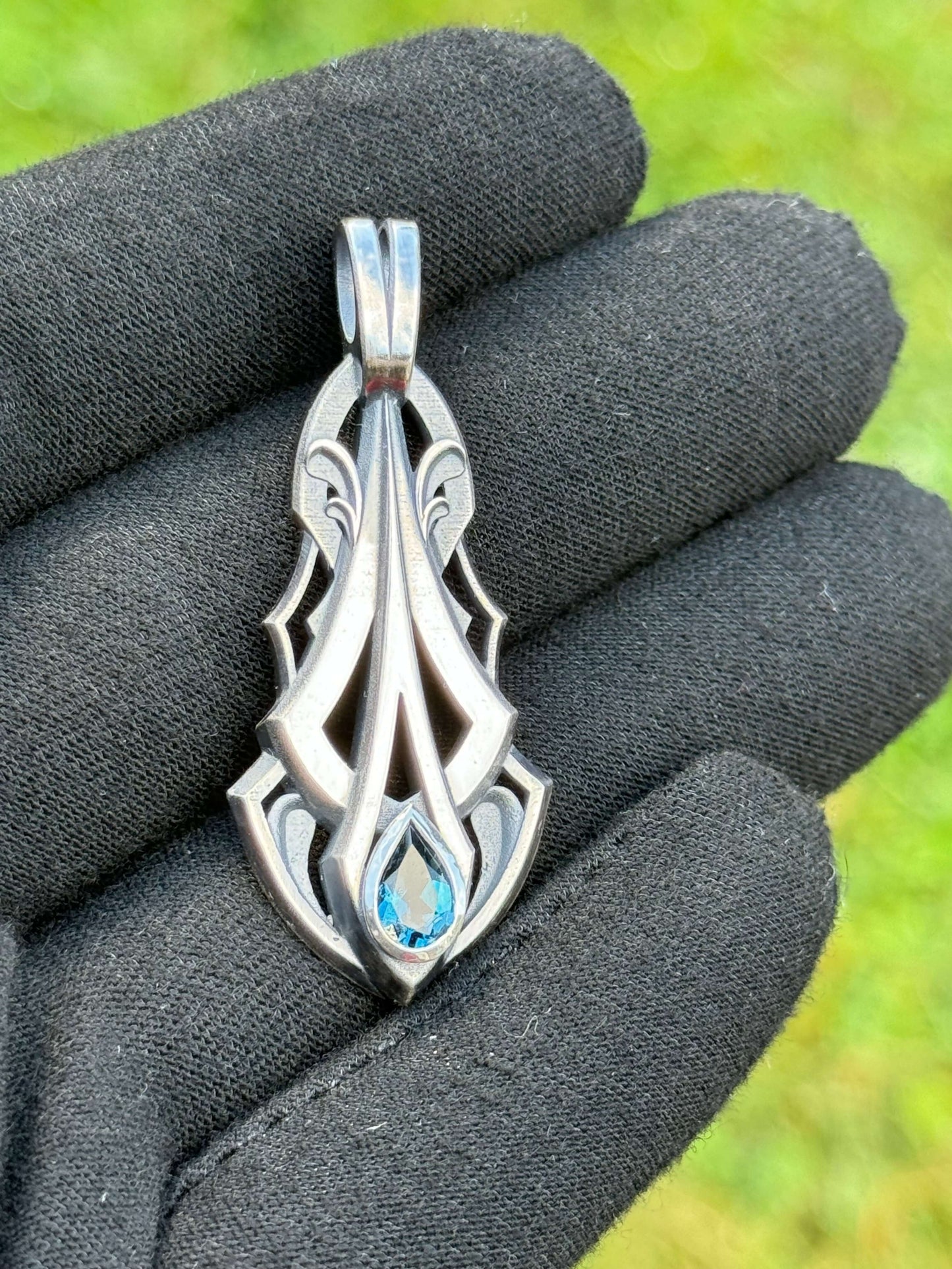 Silver pendant with an intricate design and a pear-shaped blue gemstone, held by a gloved hand against a grassy background