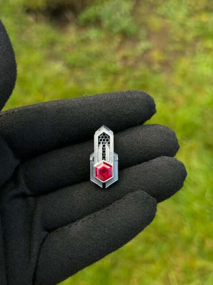 Geometric silver pendant with honeycomb details and a red gemstone, held by a gloved hand against a grassy background.