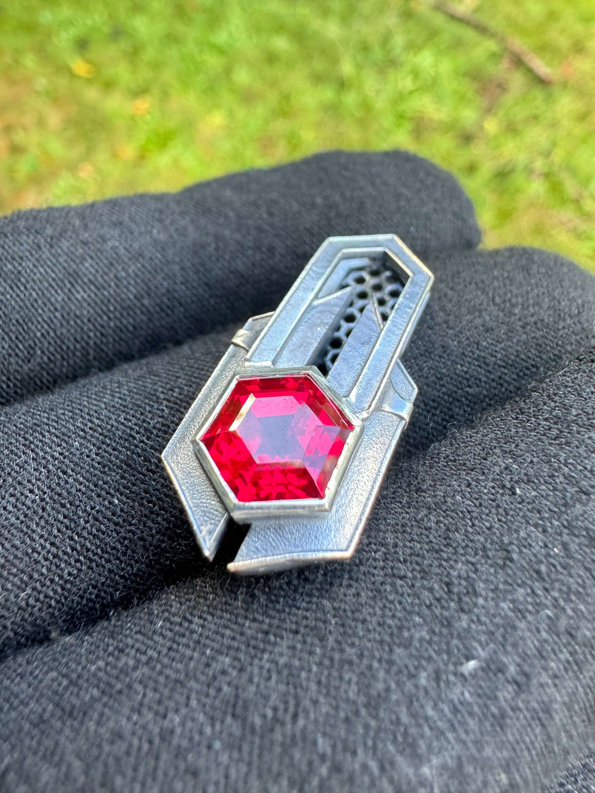 Close up of Geometric silver pendant with honeycomb details and a red gemstone, held by a gloved hand against a grassy background.