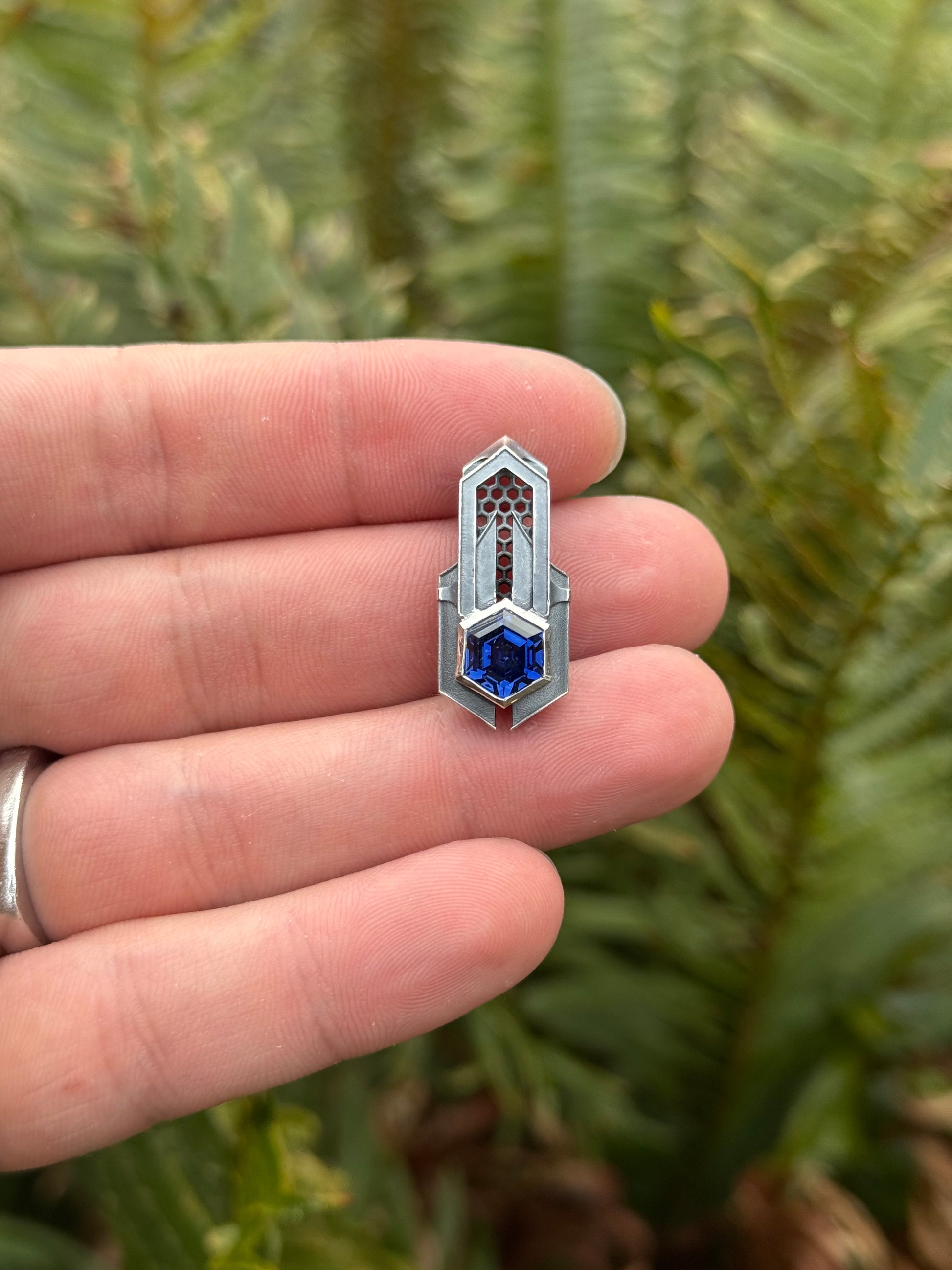 Geometric silver pendant with honeycomb details and a blue gemstone, held by a hand against a grassy background.