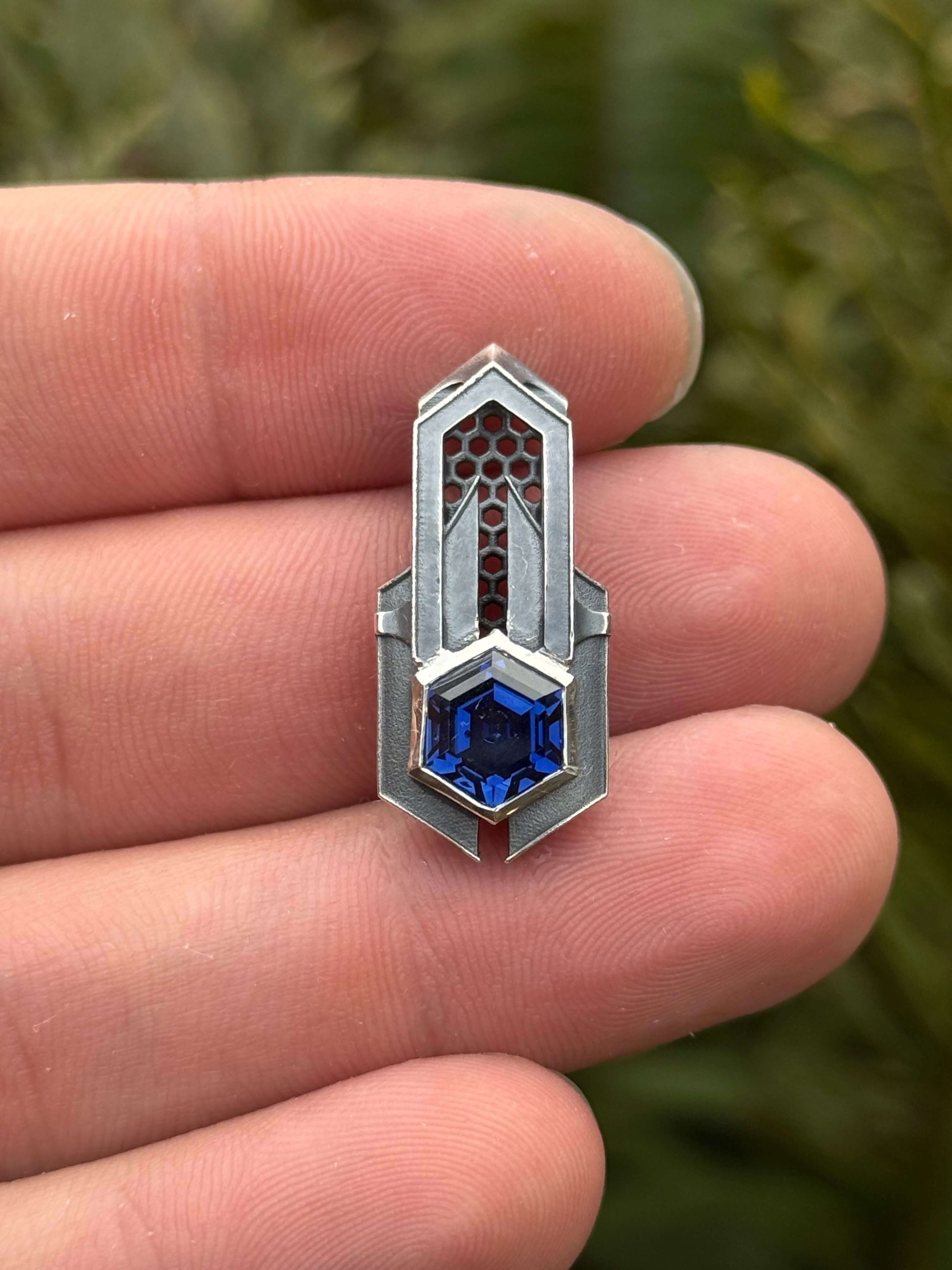 Geometric silver pendant with honeycomb details and a blue gemstone, held by a hand against a grassy background.