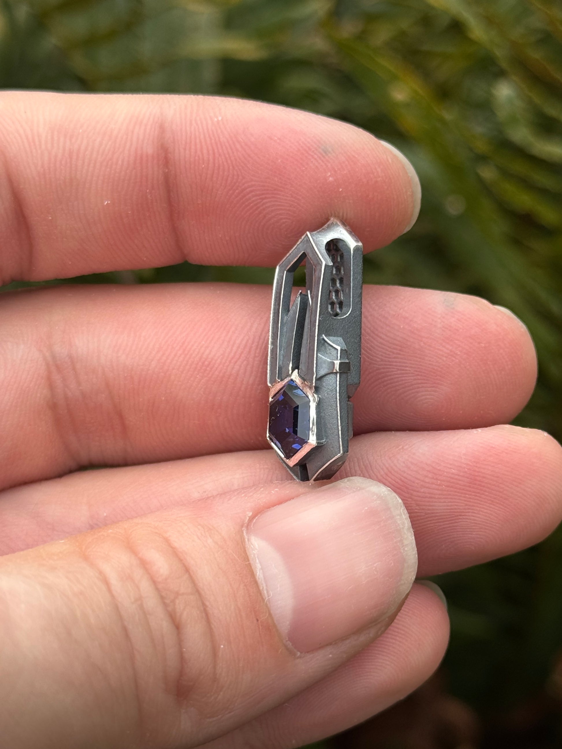 Side view of Geometric silver pendant with honeycomb details and a blue gemstone, held by a hand against a grassy background.