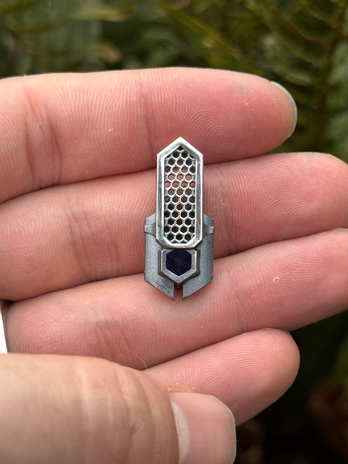 backside of Geometric silver pendant with honeycomb details and a blue gemstone, held by a hand against a grassy background.
