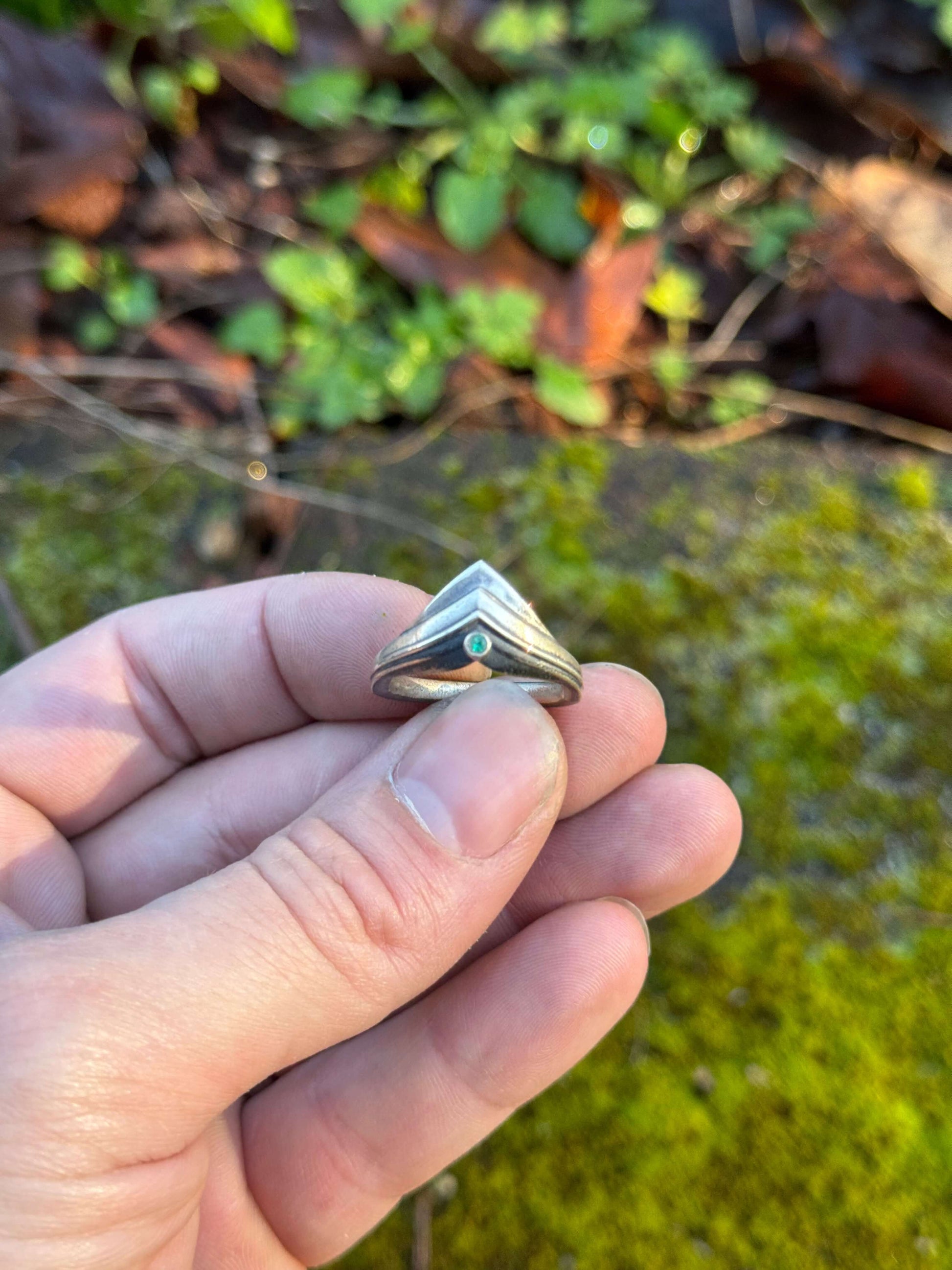 Silver ring with a modern, angular design featuring a small green gemstone, displayed in hand over vibrant moss