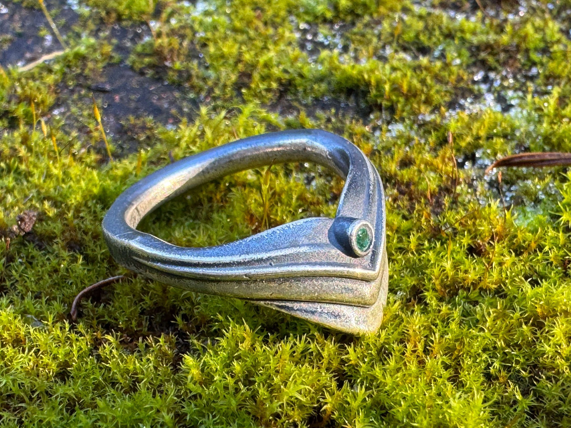 Silver ring with a modern, angular design featuring a small green gemstone, displayed on vibrant moss