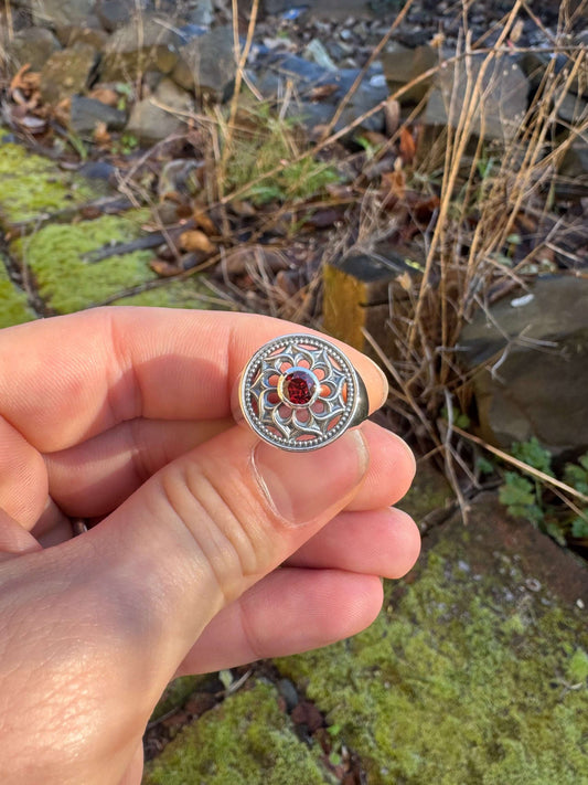 Hand holding a silver ring with a floral design and a red gemstone, set against a mossy outdoor scene with rocks and plants
