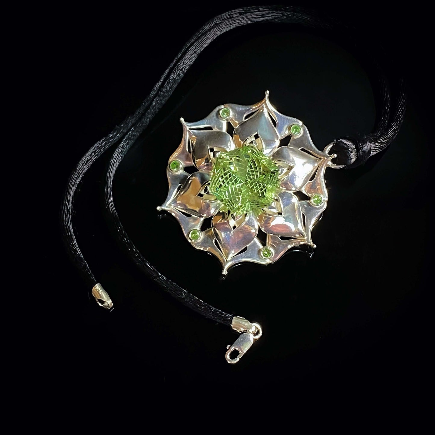 Intricate silver pendant with a floral design, featuring a green central gemstone and smaller green accents, on a black cord.