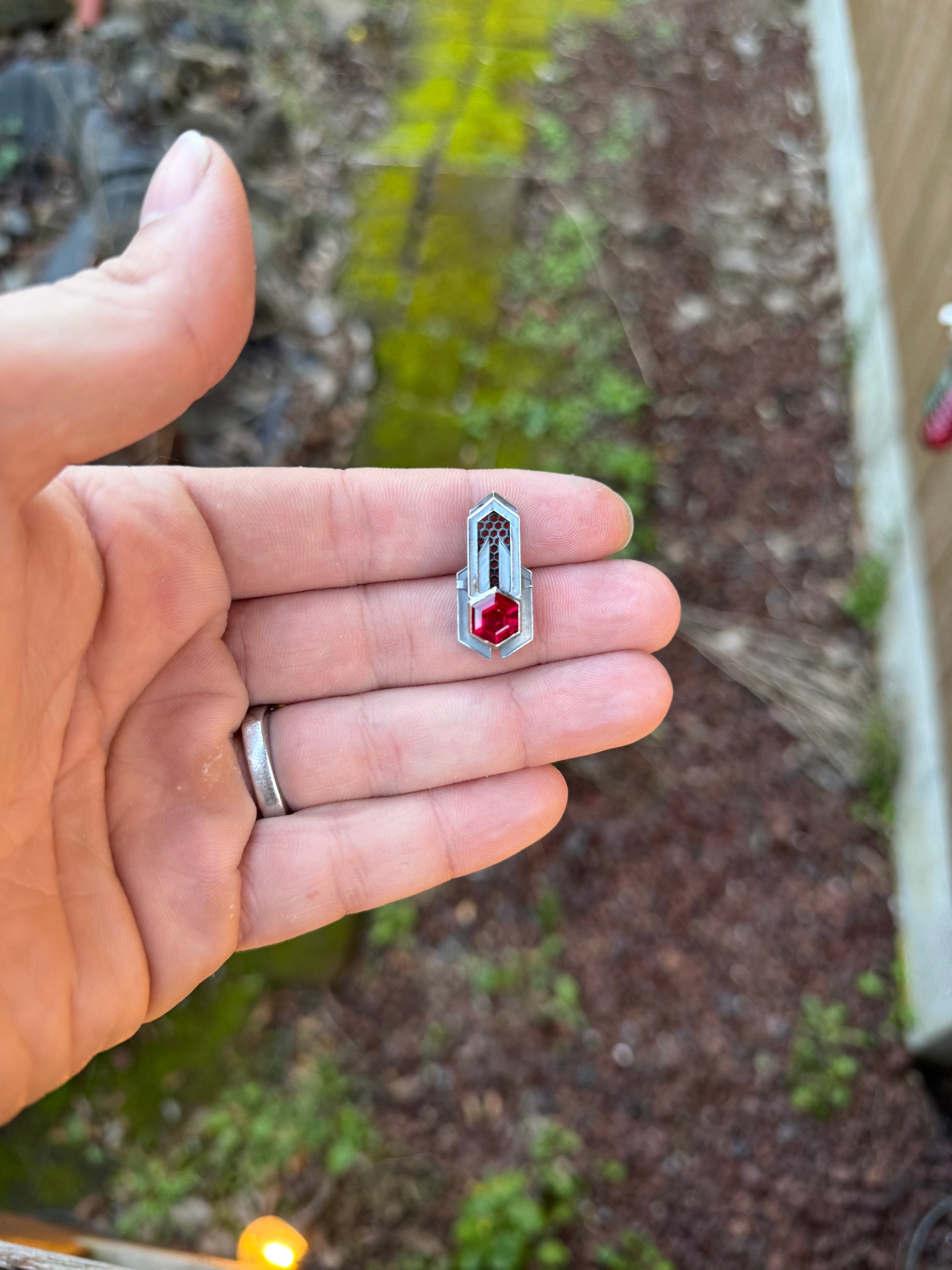 Geometric silver pendant with honeycomb details and a red gemstone, held by a hand against a grassy background.