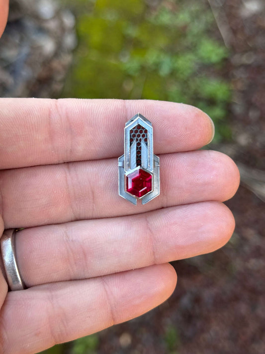 Geometric silver pendant with honeycomb details and a red gemstone, held by a hand against a grassy background.