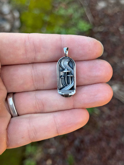 Hand holding a rectangular silver pendant featuring mushroom-shaped details and a textured background