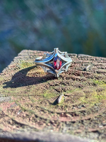 Silver ring with an ornate design featuring a marquise-cut red gemstone, displayed on a textured wooden surface