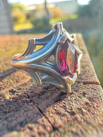 Silver ring with an ornate design featuring a large marquise-cut red gemstone, displayed on a textured wooden surface