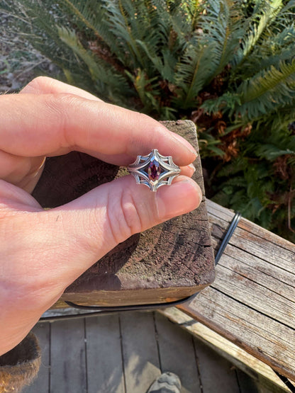 Silver ring with an ornate design featuring a large marquise-cut red gemstone, held in a hand with a fern background