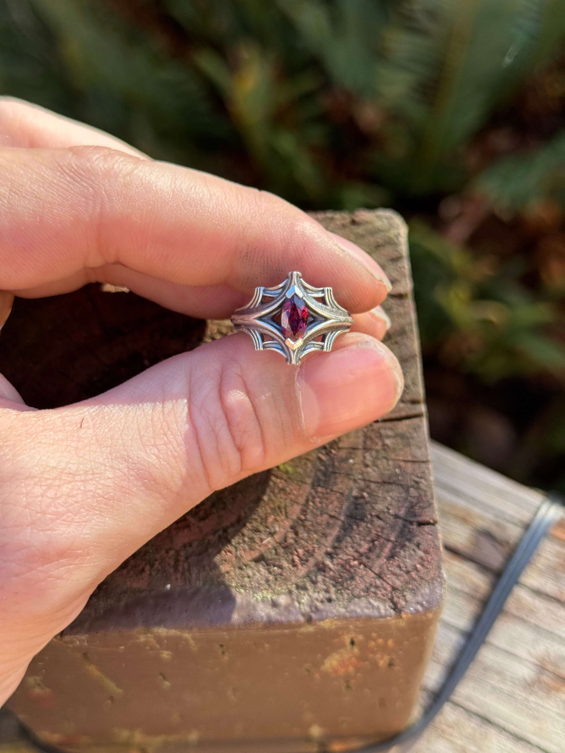 Silver ring with an ornate design featuring a large marquise-cut red gemstone, held in a hand with a fern background