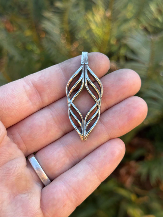 Hand holding a silver pendant with a leaf-inspired openwork design, set against a blurred natural background