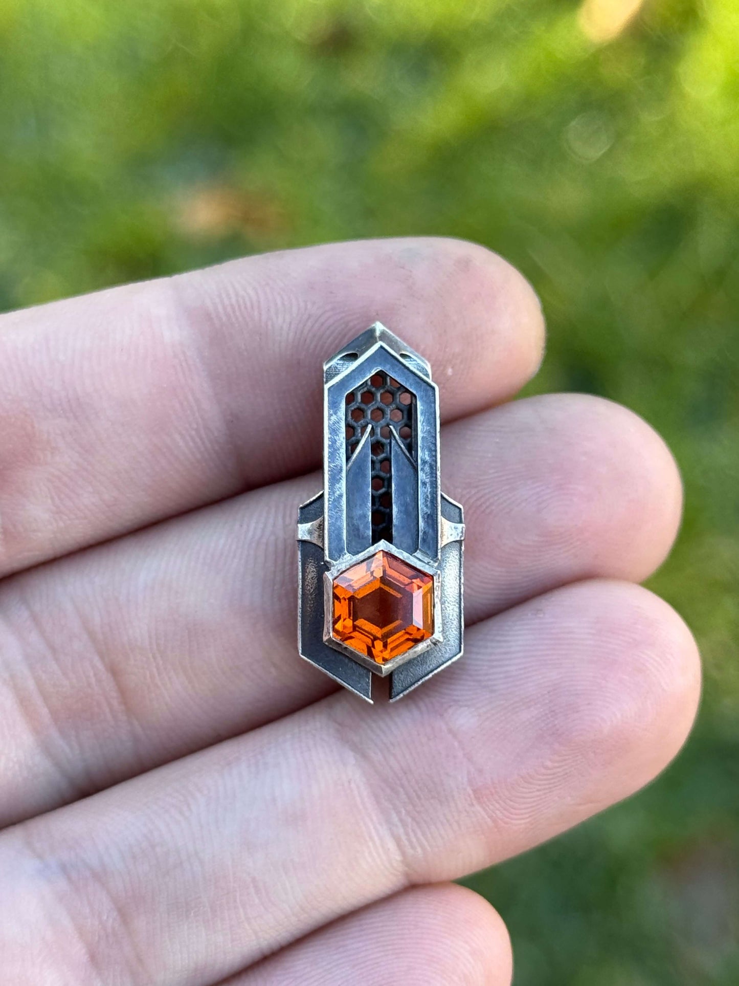 Geometric silver pendant with honeycomb details and a orange gemstone, held by a hand against a grassy background.