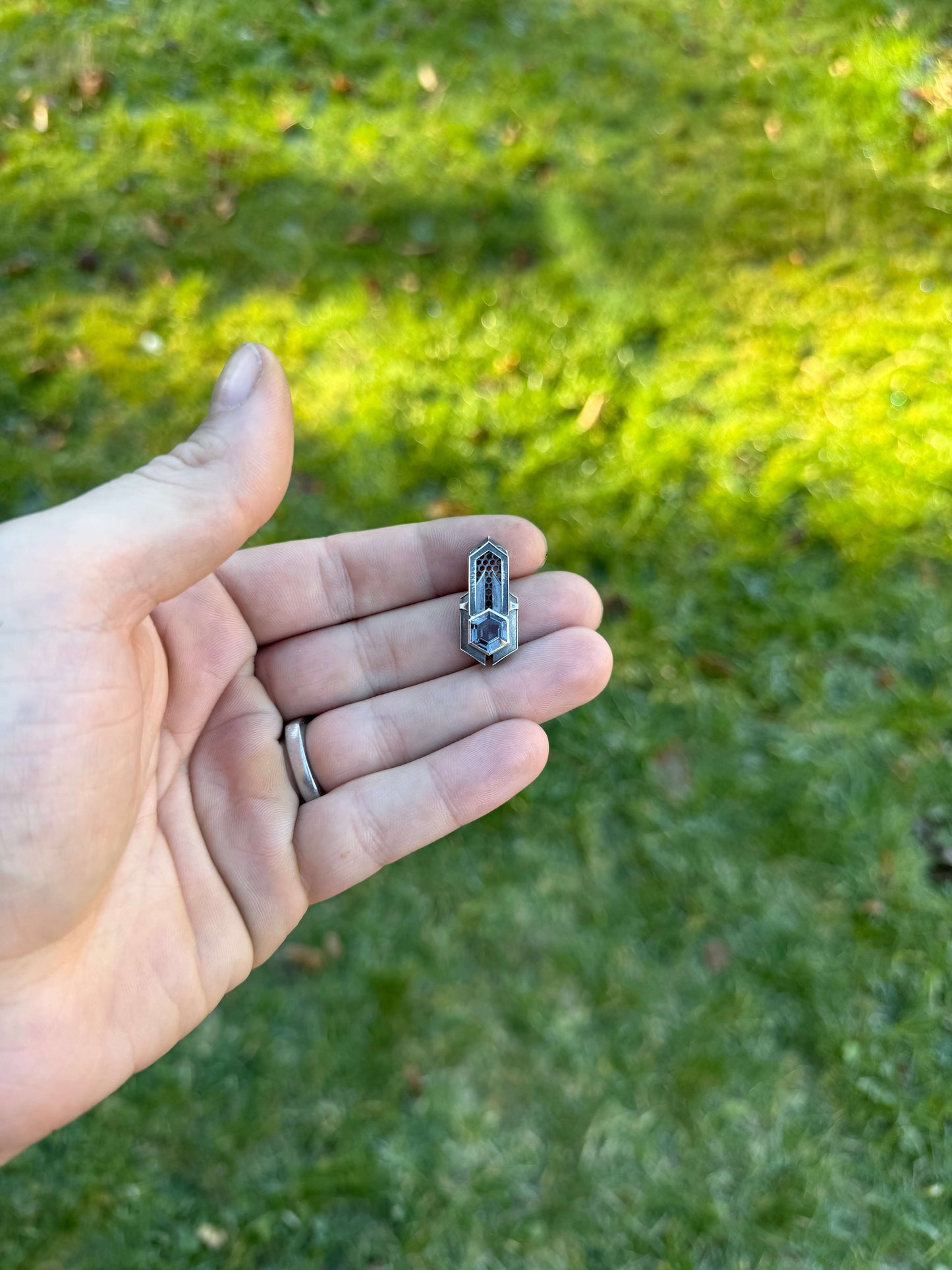 Geometric silver pendant with honeycomb details and a blue gemstone, held by a hand against a grassy background.