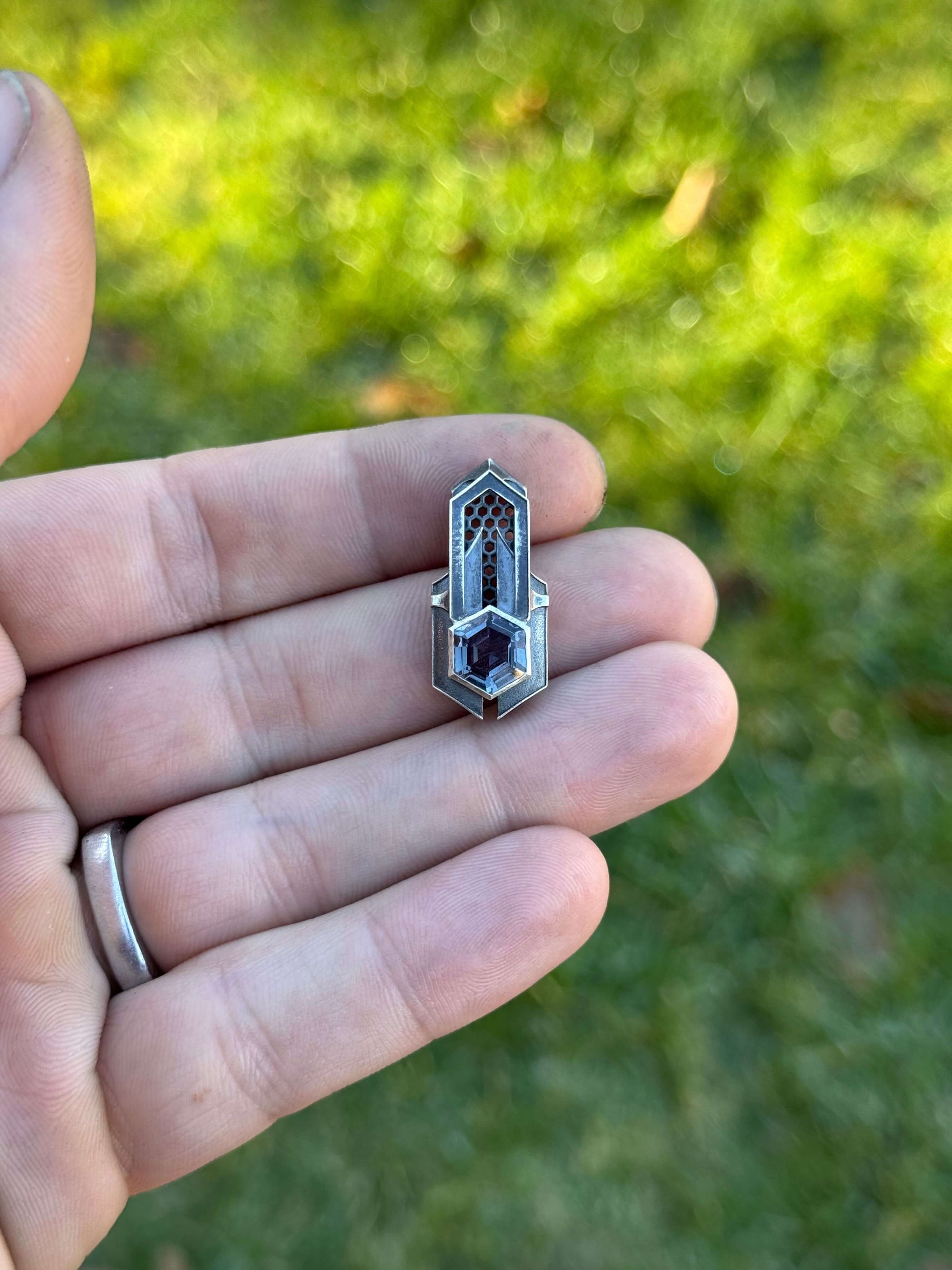 Geometric silver pendant with honeycomb details and a blue gemstone, held by a hand against a grassy background.