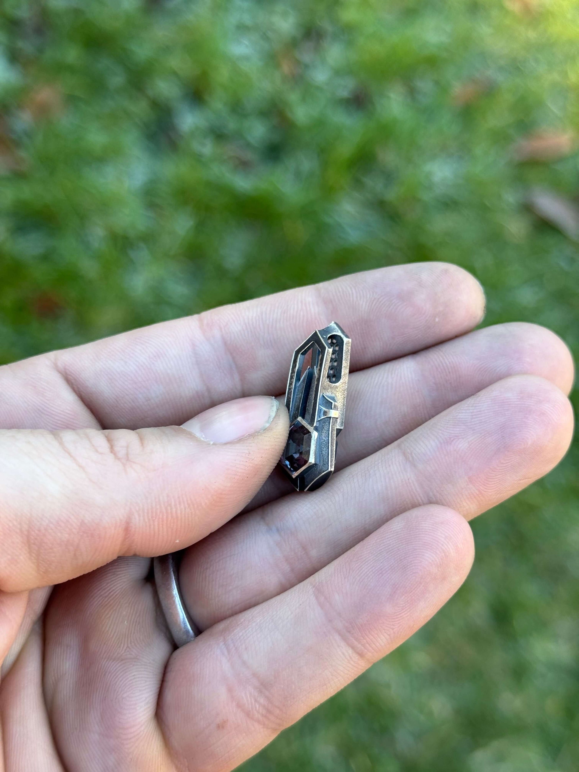 Side view of Geometric silver pendant with honeycomb details and a blue gemstone, held by a hand against a grassy background.