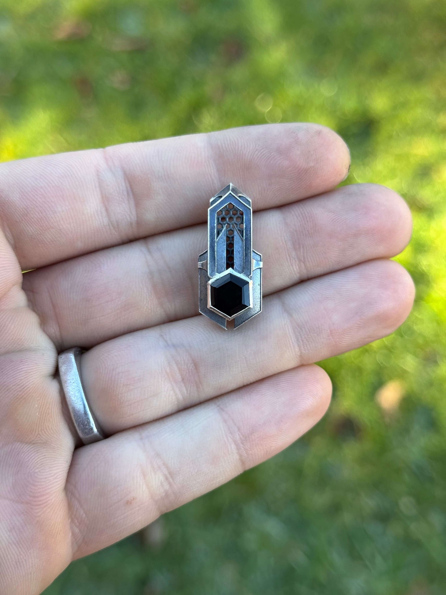 Geometric silver pendant with honeycomb details and a blue gemstone, held by a hand against a grassy background.