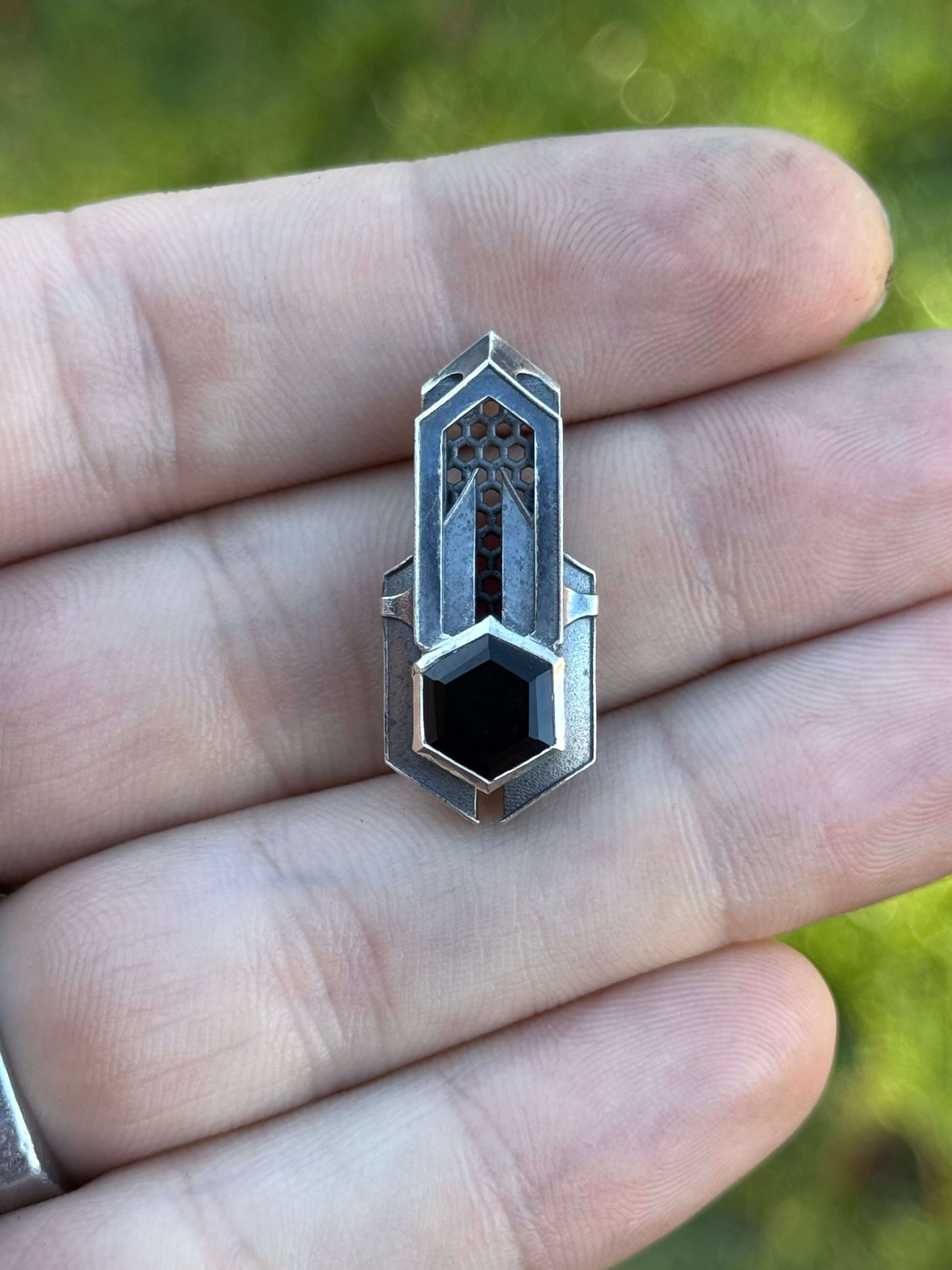 Geometric silver pendant with honeycomb details and a black gemstone, held by a hand against a grassy background.