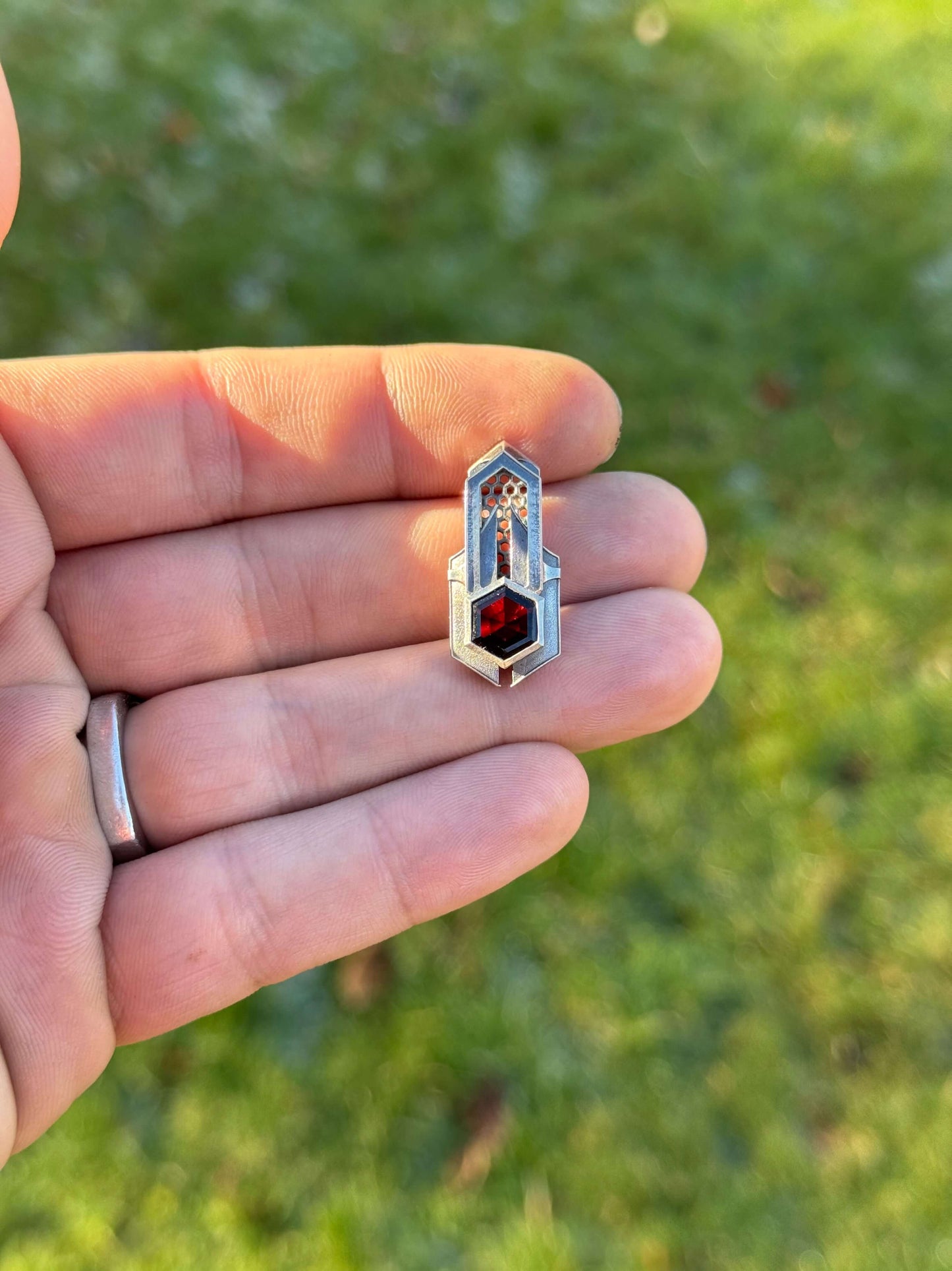 Geometric silver pendant with honeycomb details and a blue gemstone, held by a hand against a grassy background.