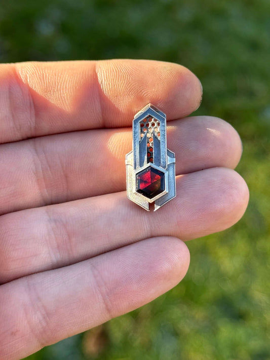 Geometric silver pendant with honeycomb details and a red gemstone, held by a hand against a grassy background.