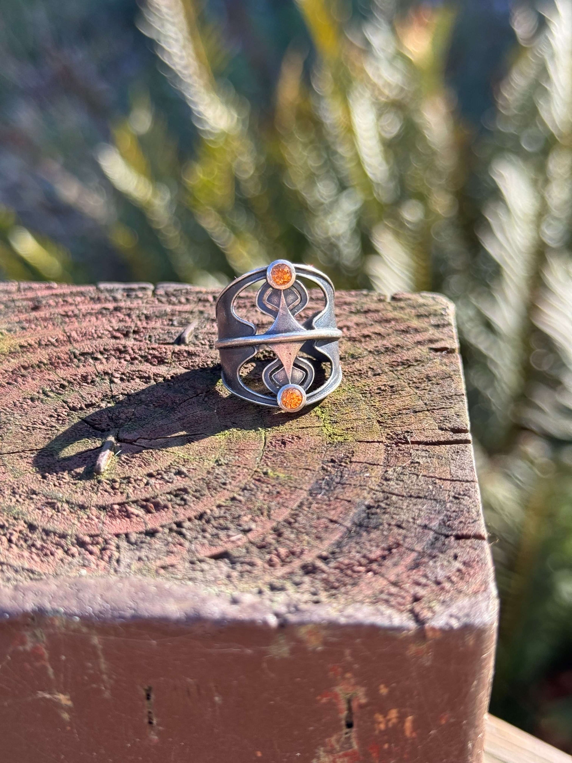 Silver ring with an abstract design featuring two orange gemstones, displayed on a textured wooden surface outdoors.