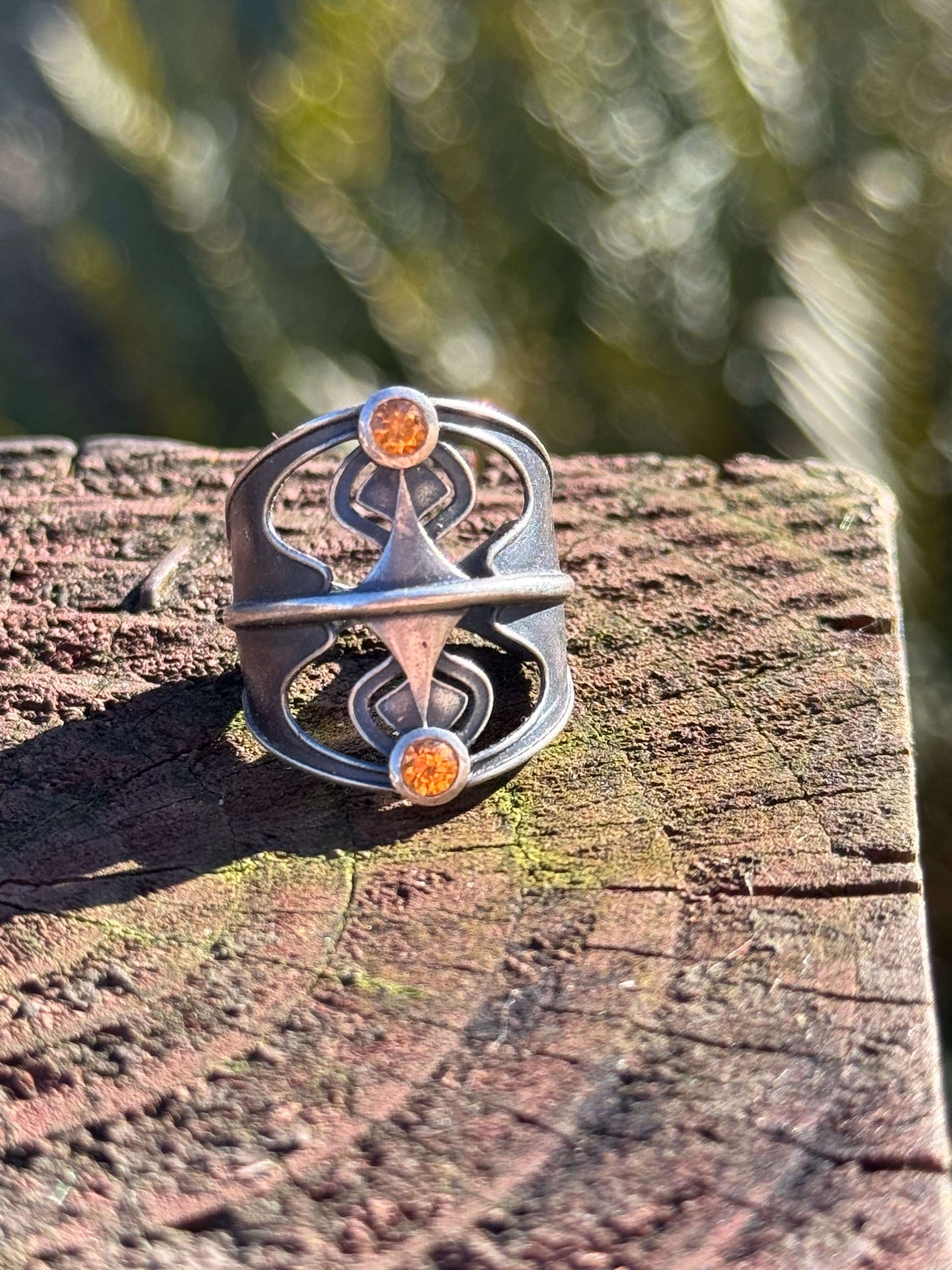 Silver ring with an abstract design featuring two orange gemstones, displayed on a textured wooden surface outdoors.