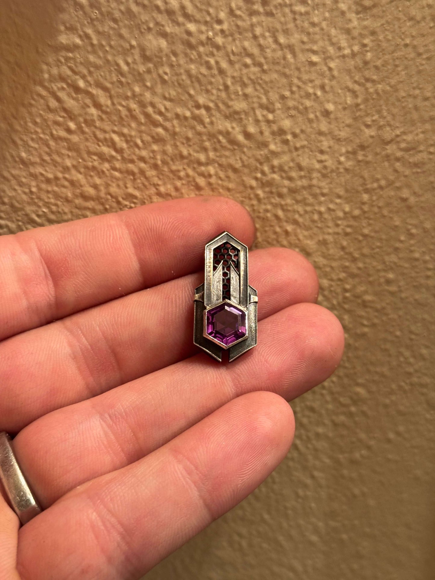 Geometric silver pendant with honeycomb details and a purple gemstone, held by a hand indoors.