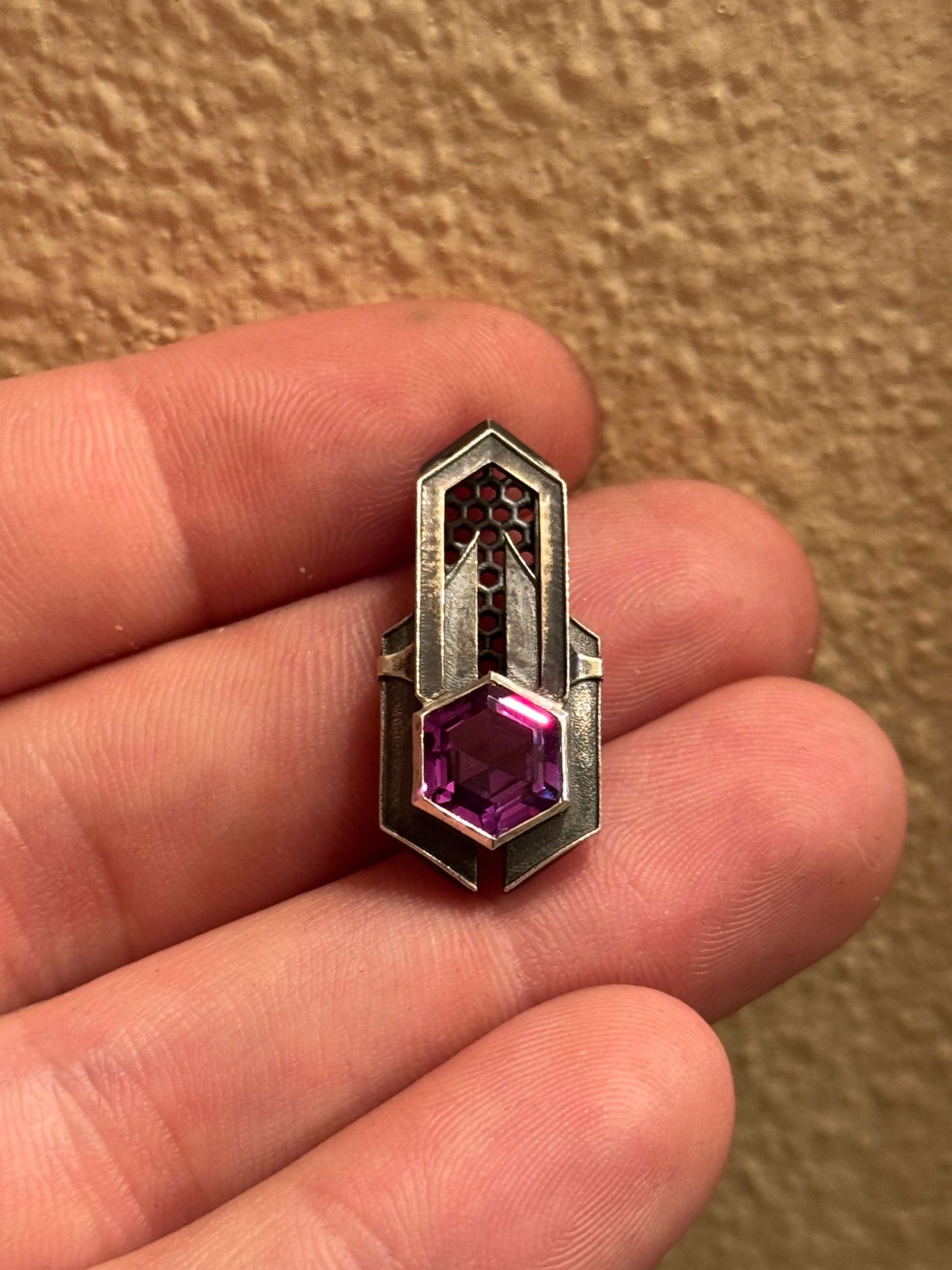 Geometric silver pendant with honeycomb details and a purple gemstone, held by a hand indoors.