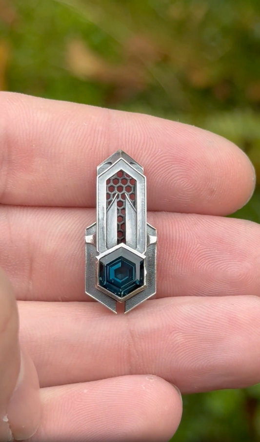 Geometric silver pendant with honeycomb details and a blue gemstone, held by a hand against a grassy background.