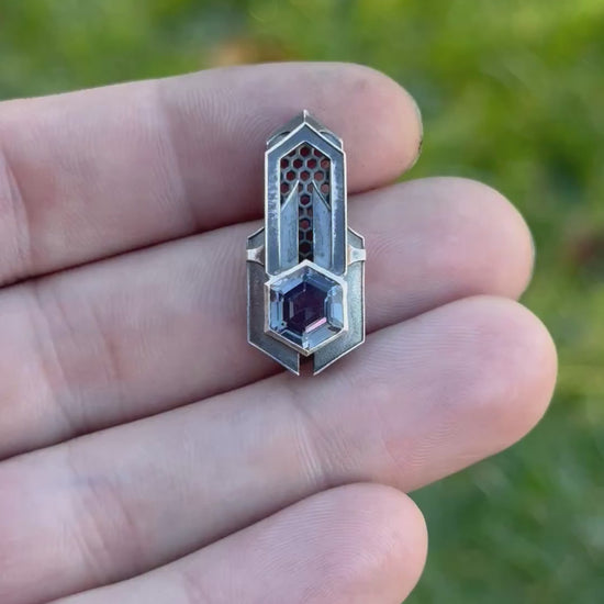 Geometric silver pendant with honeycomb details and a purple gemstone, held by a hand against a grassy background.