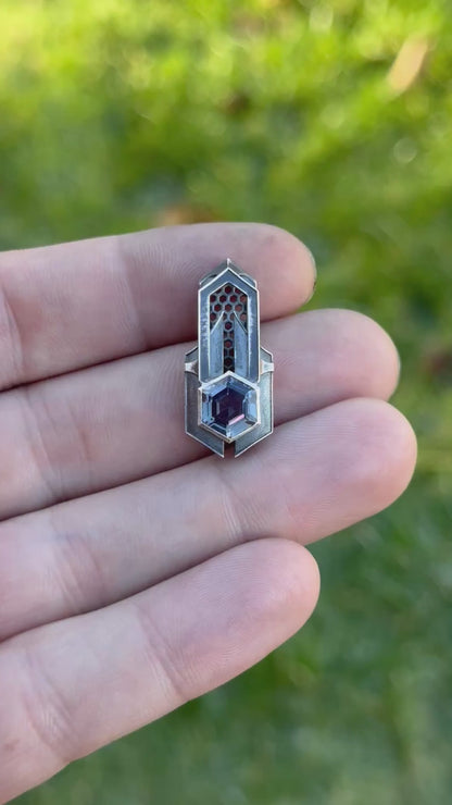 Geometric silver pendant with honeycomb details and a purple gemstone, held by a hand against a grassy background.