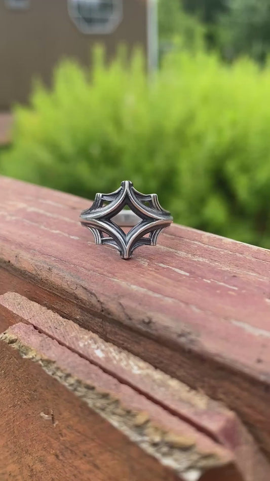Silver ring with an intricate geometric design, displayed on a wooden surface with a blurred green background