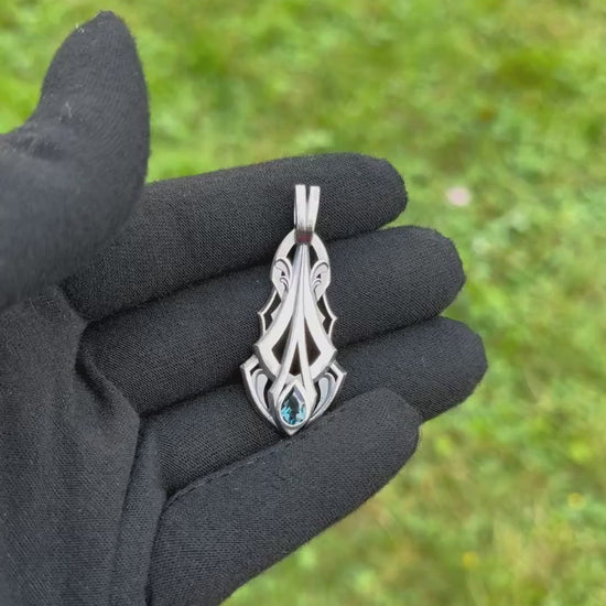 Silver pendant with an intricate design and a pear-shaped blue gemstone, held by a gloved hand against a grassy background