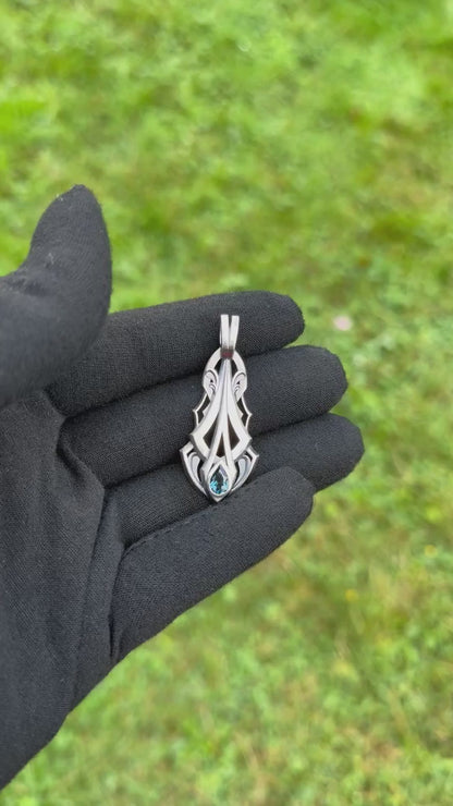 Silver pendant with an intricate design and a pear-shaped blue gemstone, held by a gloved hand against a grassy background