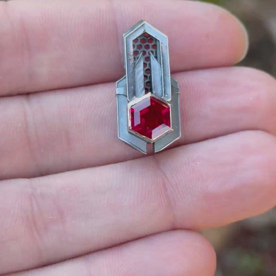 Geometric silver pendant with honeycomb details and a red gemstone, held by a hand against a grassy background.